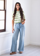 A woman stands in a bright room wearing a light green and white striped short-sleeve top, light-wash wide-leg blue jeans, and brown shoes.