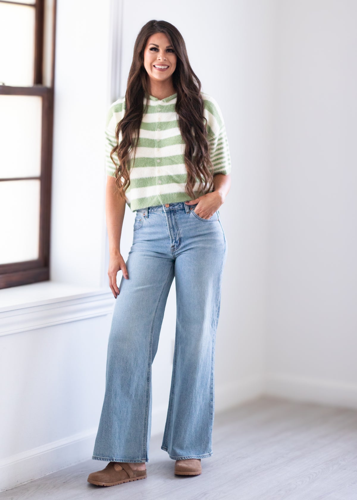 A woman stands in a bright room wearing a light green and white striped short-sleeve top, light-wash wide-leg blue jeans, and brown shoes.