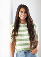 A smiling woman with long wavy brown hair wearing a fuzzy short-sleeve button-front cardigan with horizontal sage green and cream stripes, paired with light-wash blue jeans.