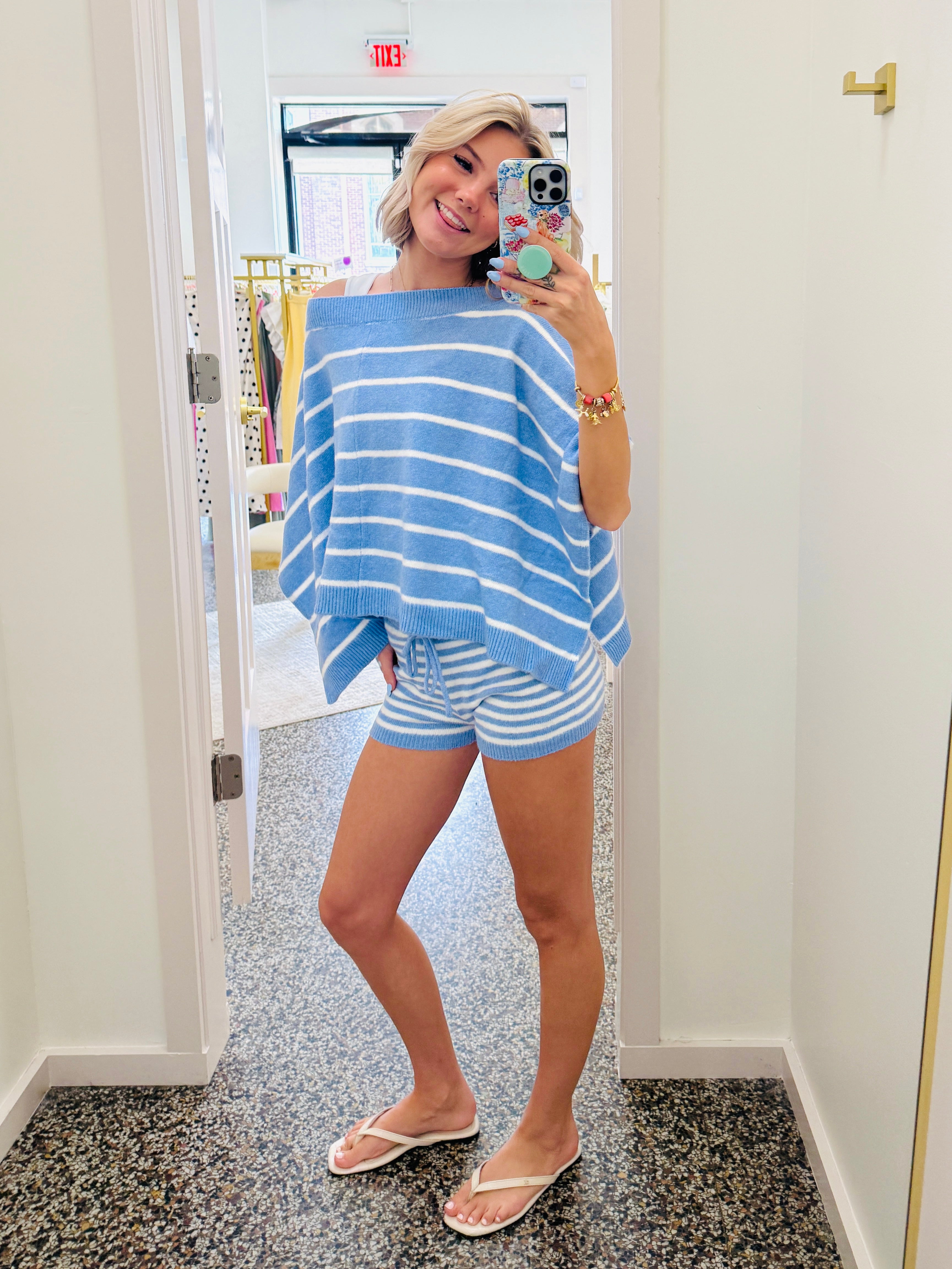 A woman takes a mirror selfie in a fitting room wearing a light blue and white striped poncho-style top and matching striped shorts.