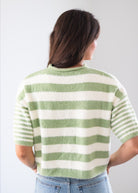 A woman viewed from behind wearing a fuzzy short-sleeve knit sweater with horizontal sage green and off-white stripes, paired with light blue jeans against a plain light gray background.