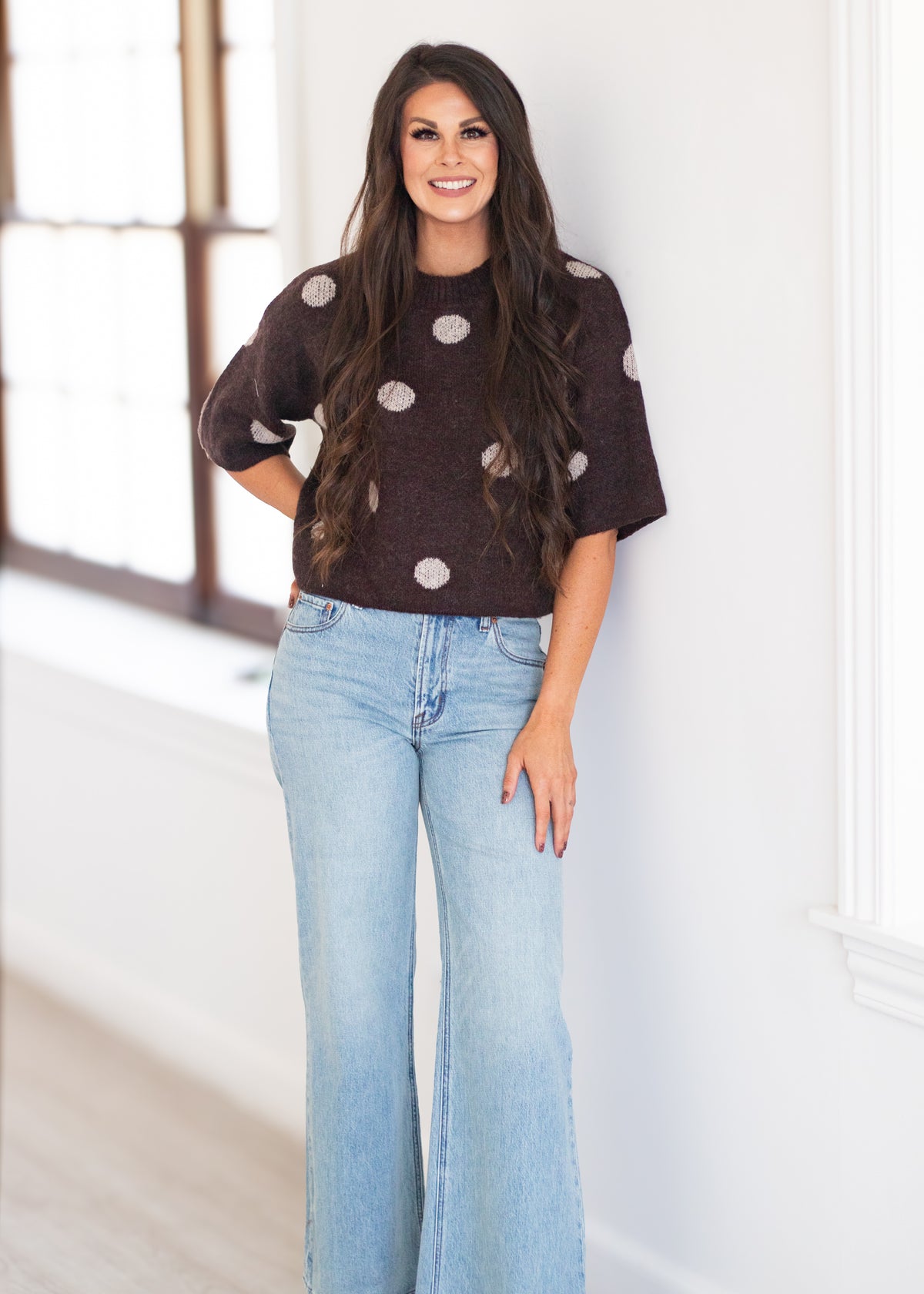 A woman stands indoors wearing a dark brown short-sleeve knit sweater with large light beige polka dots and light-wash blue flare jeans.