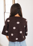 A woman viewed from behind wearing a dark brown knit sweater with large white polka dots and light blue jeans, standing indoors against a bright white wall.