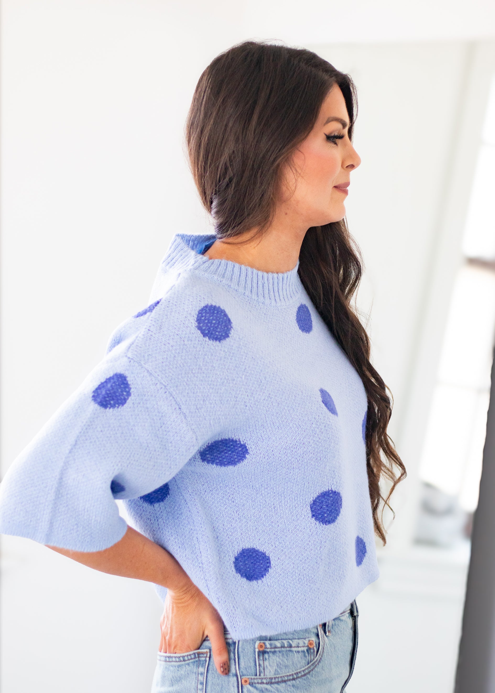 A woman wearing a light blue knit sweater with dark blue polka dots, paired with light-wash jeans, shown in a bright indoor setting.