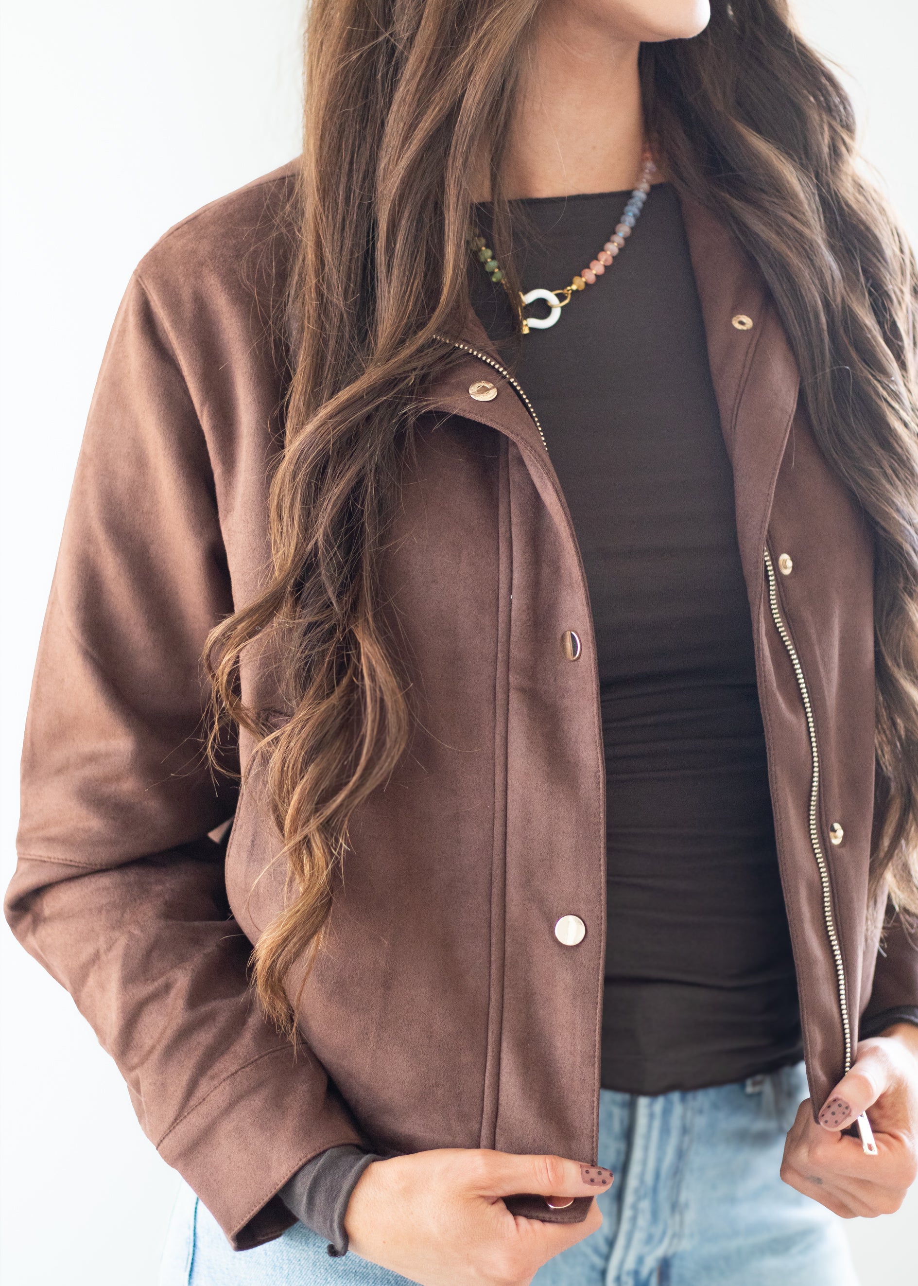 A woman wearing a brown suede-like jacket over a black top and light blue jeans, shown from the neck down against a plain white background.