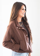 A woman wearing a brown cropped jacket with snap buttons and a pointed collar, styled over a dark top and light blue jeans, photographed against a plain white background.