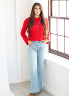 A woman stands indoors next to a window wearing a bright red long-sleeve knit sweater, light-wash blue flare jeans, and a colorful beaded necklace.