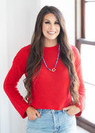 A smiling woman with long brown hair wearing a bright red knit sweater and light blue jeans, standing indoors near a window.
