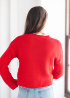 A woman viewed from behind wearing a bright red knit sweater and light blue jeans, standing in a brightly lit indoor setting.