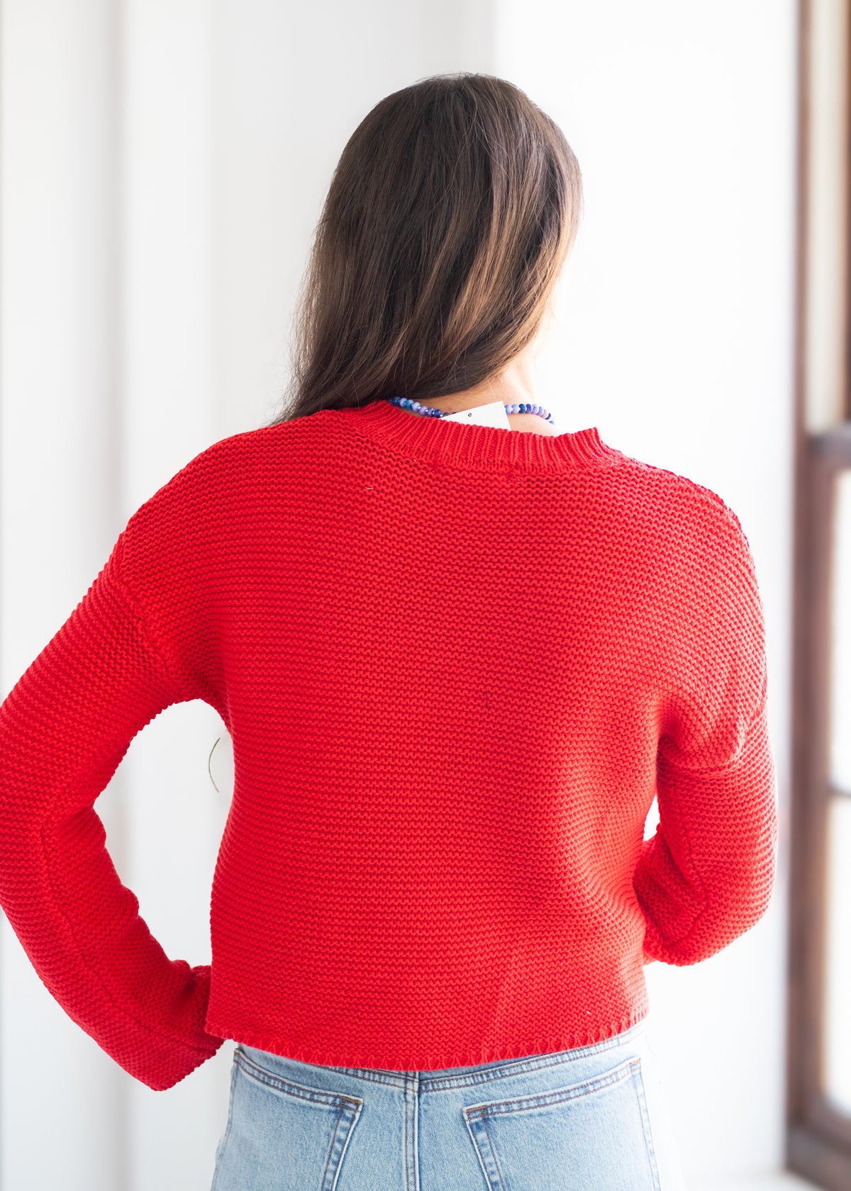 A woman viewed from behind wearing a bright red knit sweater and light blue jeans, standing in a brightly lit indoor setting.