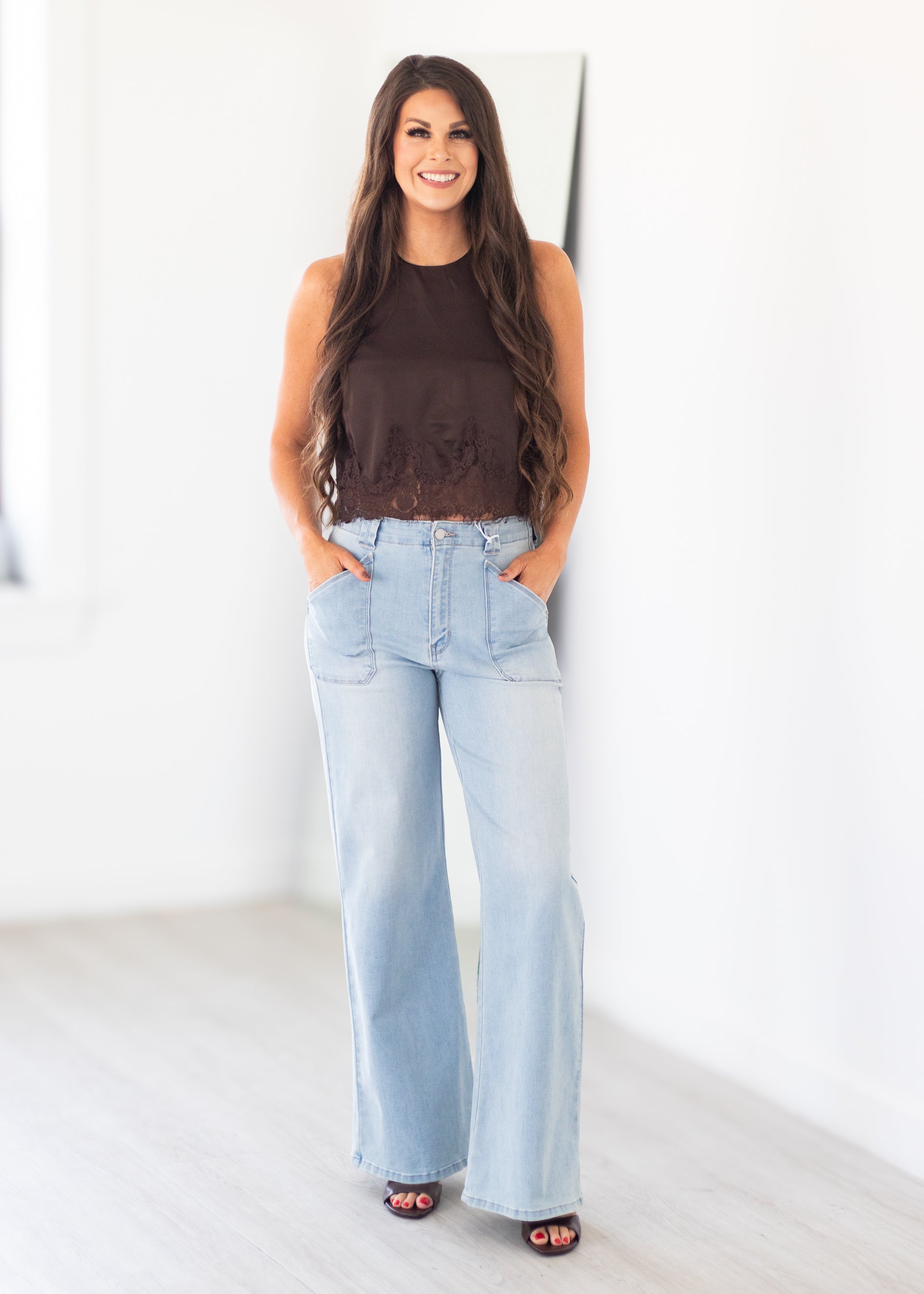 A woman stands in a bright white room wearing a sleeveless dark brown top and light-wash wide-leg denim jeans with her hands in her pockets.