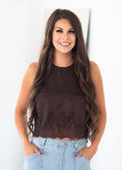 A smiling woman with long wavy brown hair wearing a dark brown sleeveless top with a lace hem and light blue jeans, standing against a plain white wall.