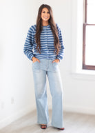 A woman stands in a bright, minimalist room wearing a blue and white striped long-sleeve button-up top with light-wash wide-leg jeans.