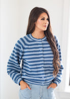 A woman wearing a blue and light blue horizontally striped knit cardigan sweater with white buttons, paired with light-wash jeans, standing against a bright white indoor background.