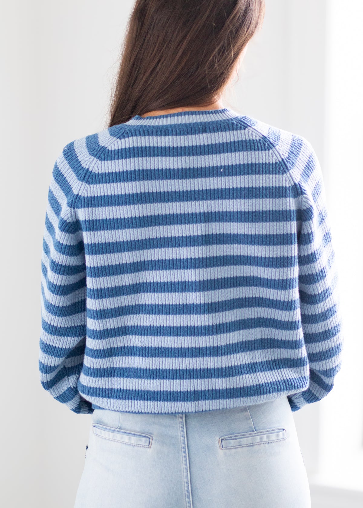 A person shown from the back wearing a blue and white horizontally striped knit sweater with light blue jeans against a bright white indoor background.