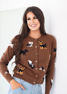 A woman wearing a brown button-up cardigan sweater with a playful pattern of various dog and cat illustrations, paired with light blue jeans, posing against a plain white wall.