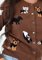 A close-up of a brown knit cardigan sweater featuring knitted dog illustrations in black, white, and tan, with silver buttons on the front. A person wearing it is partially visible, with long brown hair and light blue jeans also shown.