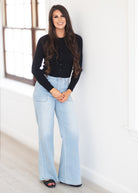 A woman stands indoors wearing a black long-sleeve button-front top and light-wash wide-leg jeans, with black sandals, against a bright white wall near a window.