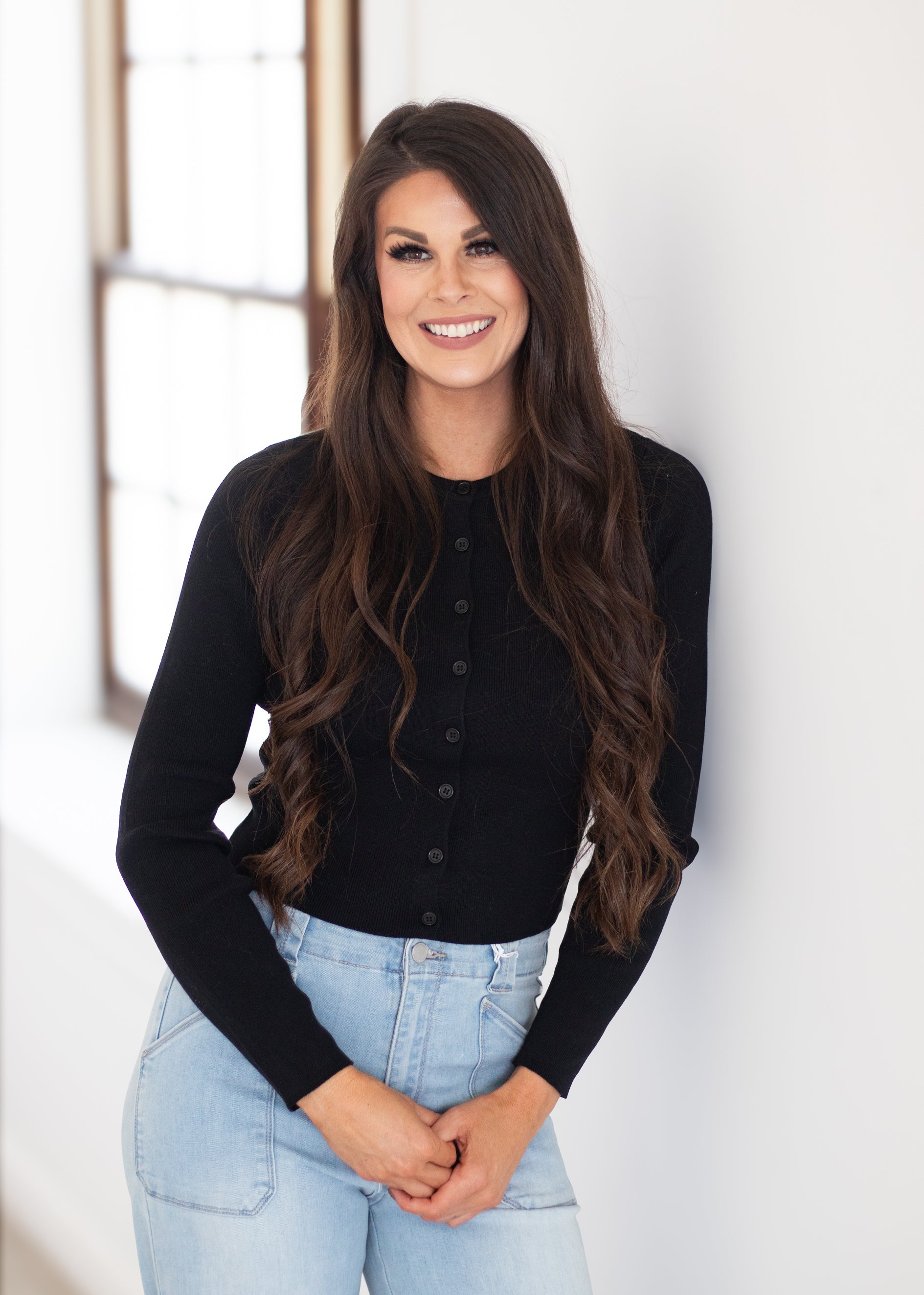 A smiling woman with long wavy brown hair stands indoors against a white wall, wearing a black long-sleeve button-front top and light blue jeans.