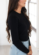 A woman wearing a black long-sleeve button-front cardigan and light blue jeans, shown in a side profile view in a bright indoor setting.