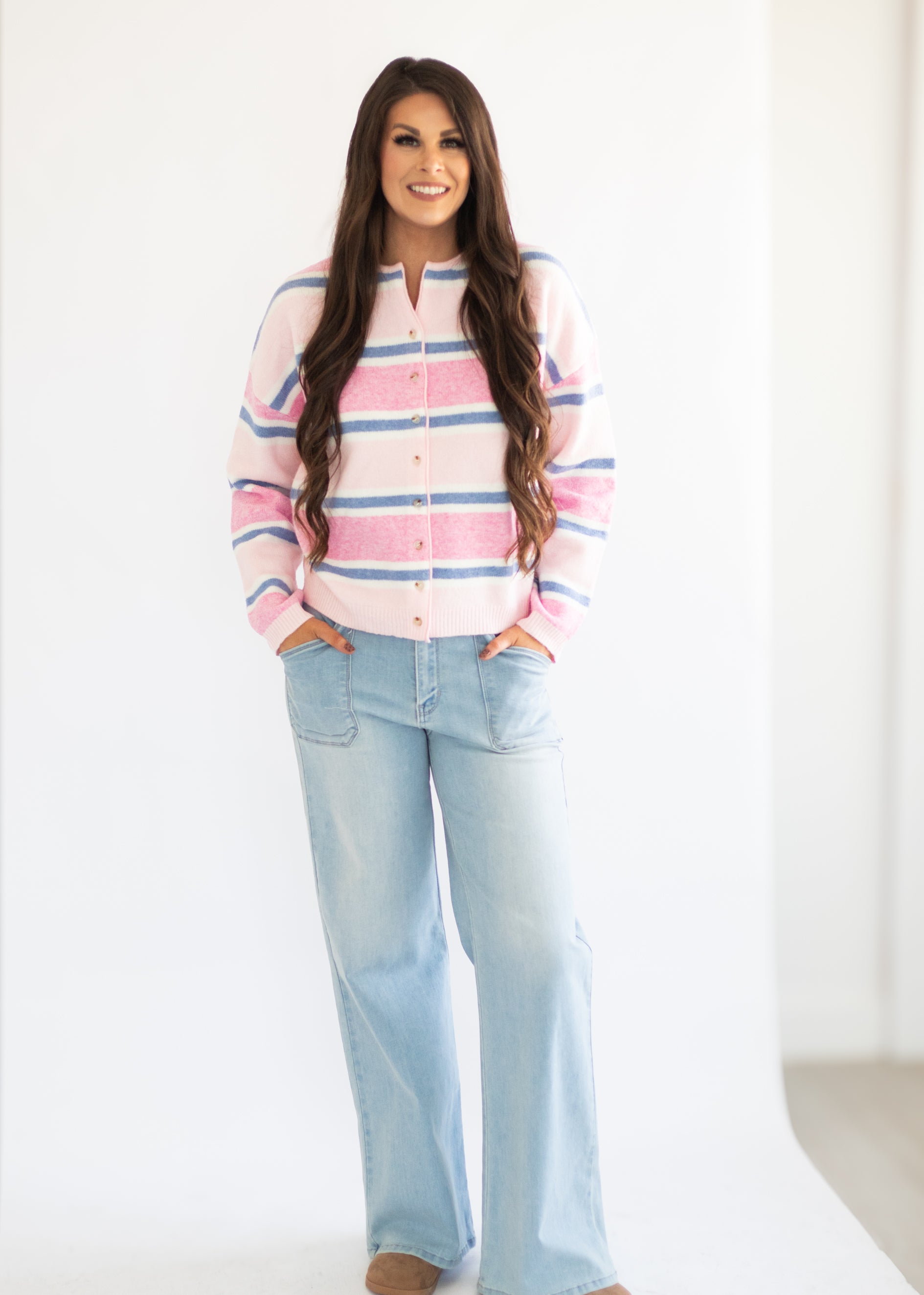 A woman modeling a pink, light blue, and white striped knit cardigan with a split neckline, paired with light-wash wide-leg jeans, against a bright white studio background.