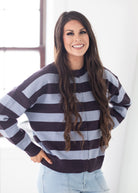 A woman with long wavy brown hair wearing a dark brown and light gray striped button-up cardigan sweater and light blue jeans, standing in a brightly lit room.