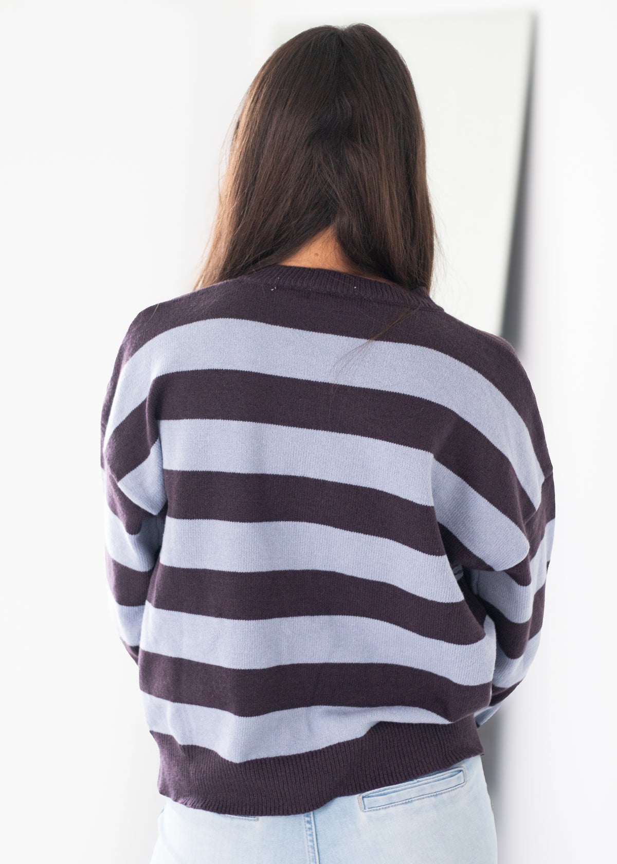 A woman viewed from behind wearing a dark brown and light gray horizontally striped sweater, paired with light blue jeans, against a plain white background.