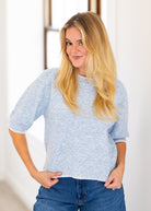 A woman with long blonde hair wearing a light blue heathered short-sleeve knit sweater and blue jeans, standing in a brightly lit indoor setting.