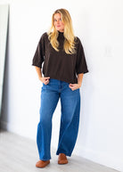 A woman stands indoors wearing a dark brown mock-neck tunic top, blue wide-leg jeans, and brown shoes against a white wall.