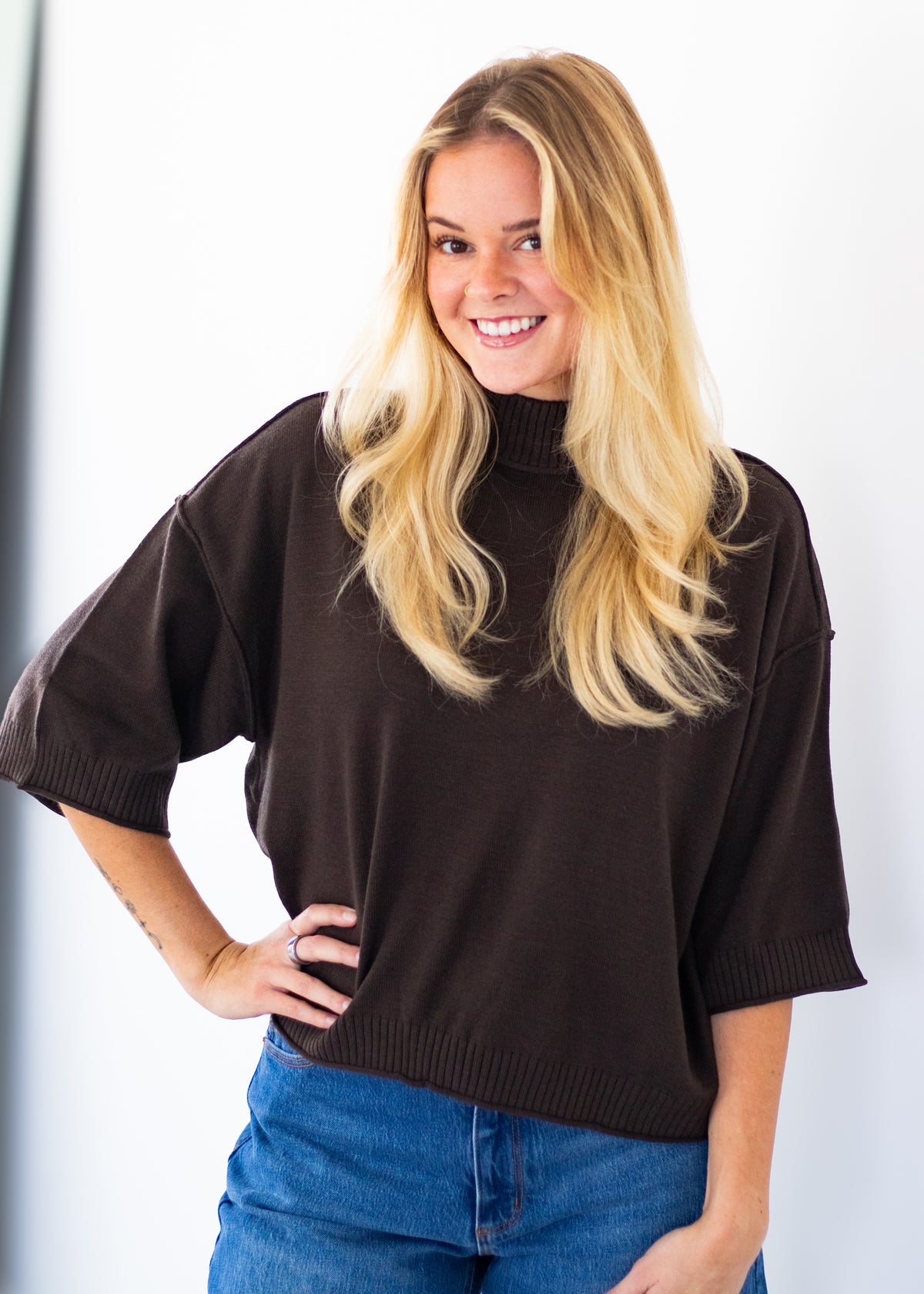 A smiling blonde woman wearing a dark brown short-sleeve mock neck knit sweater and blue jeans, standing against a plain white background.