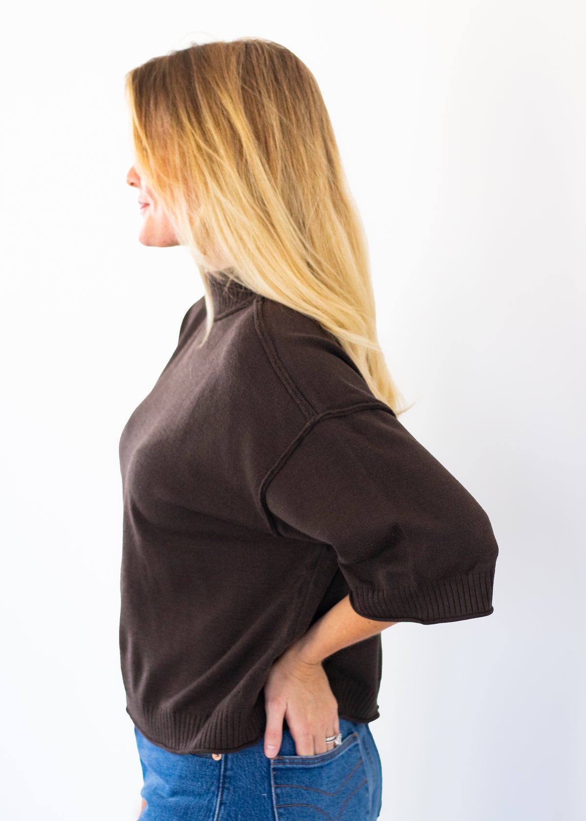 A woman with blonde hair is shown from the side profile wearing a dark brown knit mock-neck sweater and blue jeans against a plain white background.
