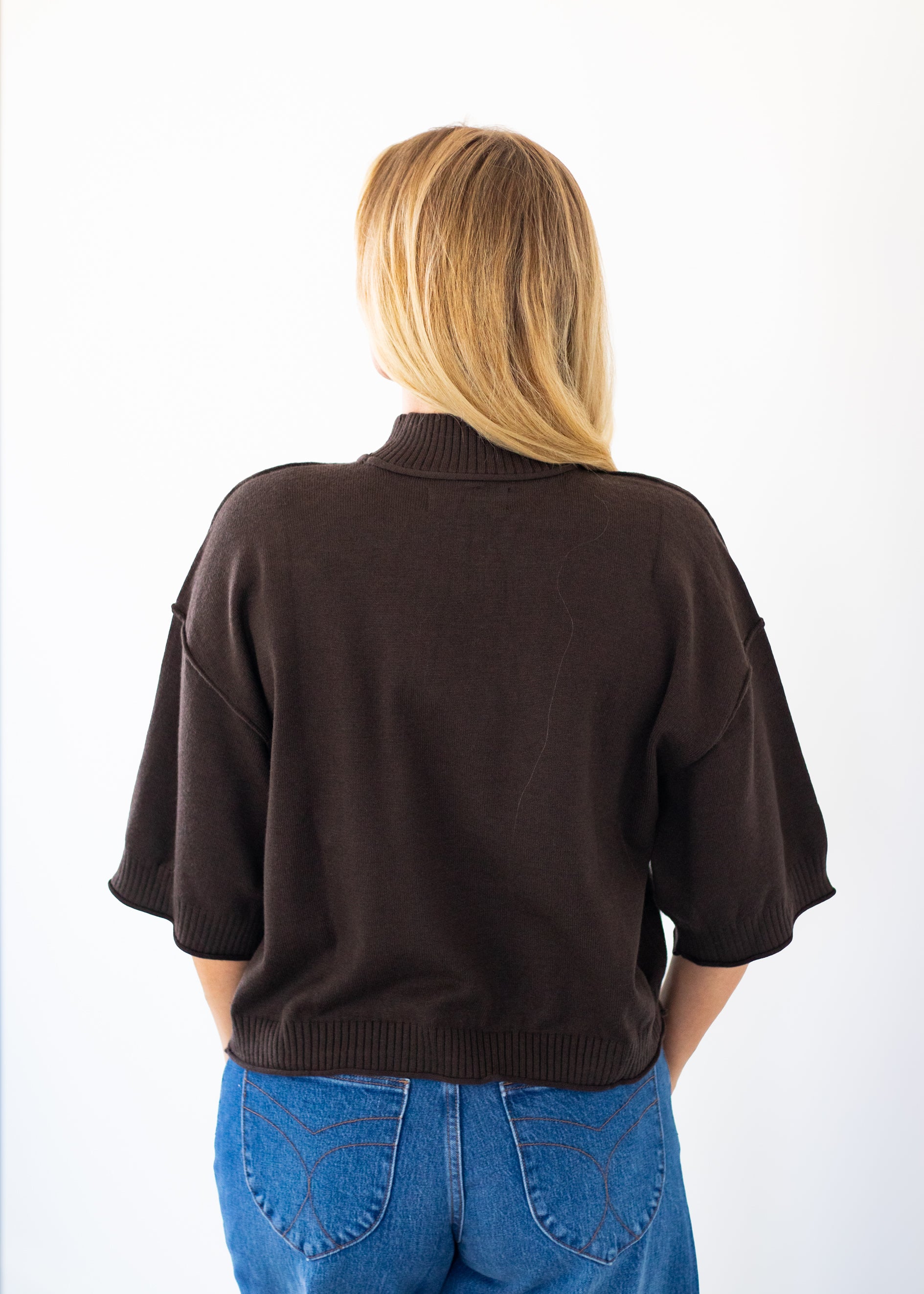 A woman viewed from behind wearing a dark brown cropped knit sweater with elbow-length sleeves and blue jeans, set against a plain white background.