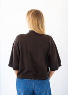 A woman viewed from behind wearing a dark brown cropped knit sweater with elbow-length sleeves and blue jeans, set against a plain white background.