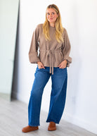 A woman modeling a taupe long-sleeve knit top with a tie waist, paired with blue wide-leg jeans and brown slip-on shoes, standing in a bright indoor space.