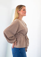 A woman wearing a light brown knit sweater with balloon sleeves and a tie waist, paired with blue jeans, shown in profile against a bright white background.