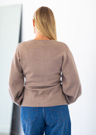 A woman viewed from behind wearing a taupe-brown long-sleeve knit sweater and blue jeans, standing against a bright white wall.