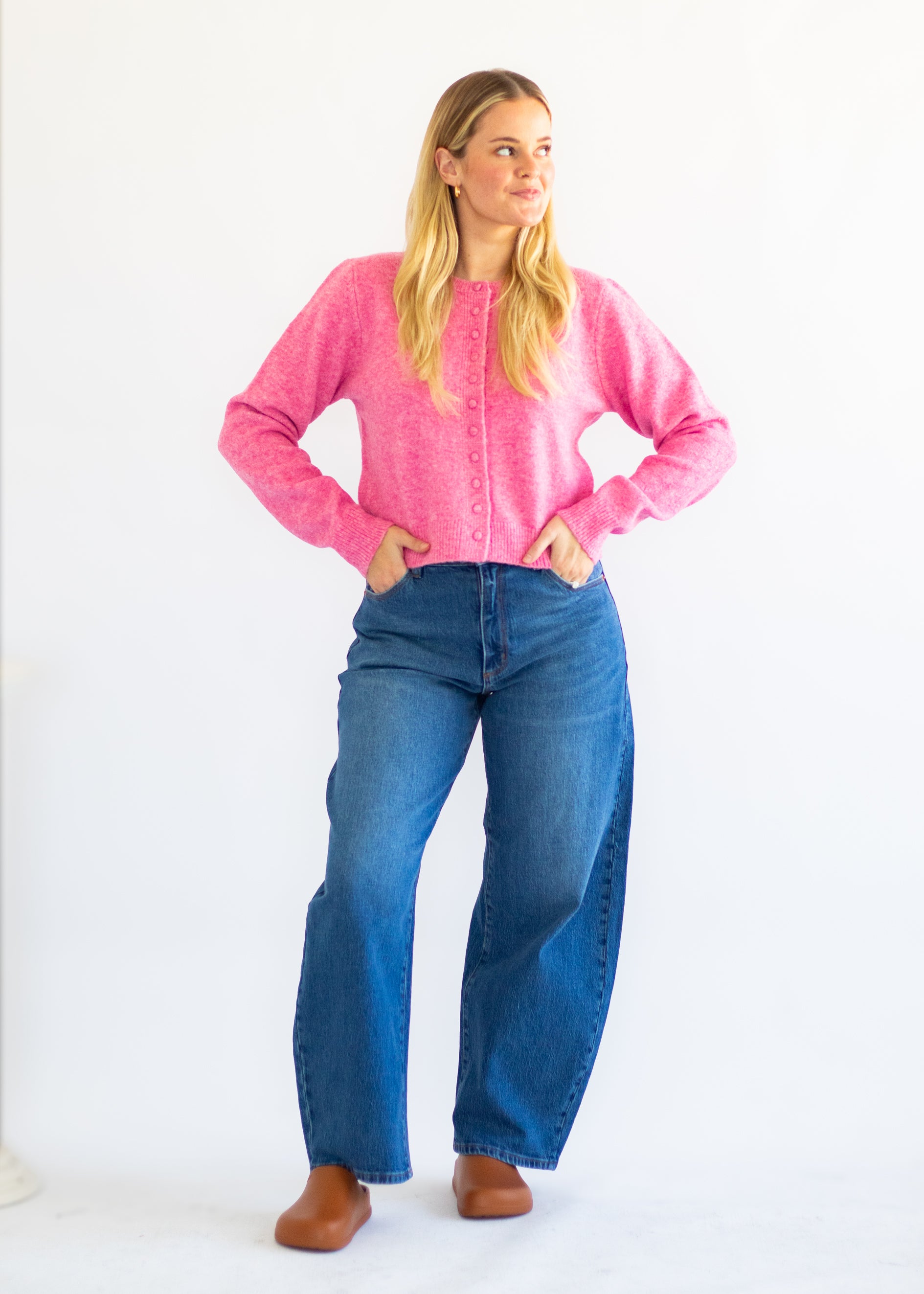 A woman modeling a bright pink long-sleeve knit cardigan with buttons, paired with high-waisted blue jeans and brown shoes, standing against a plain white studio background.