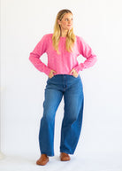A woman modeling a bright pink long-sleeve knit cardigan with buttons, paired with high-waisted blue jeans and brown shoes, standing against a plain white studio background.