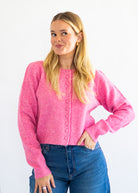 A blonde woman wearing a bright pink knit cardigan sweater with front buttons and blue jeans, standing against a plain white background.
