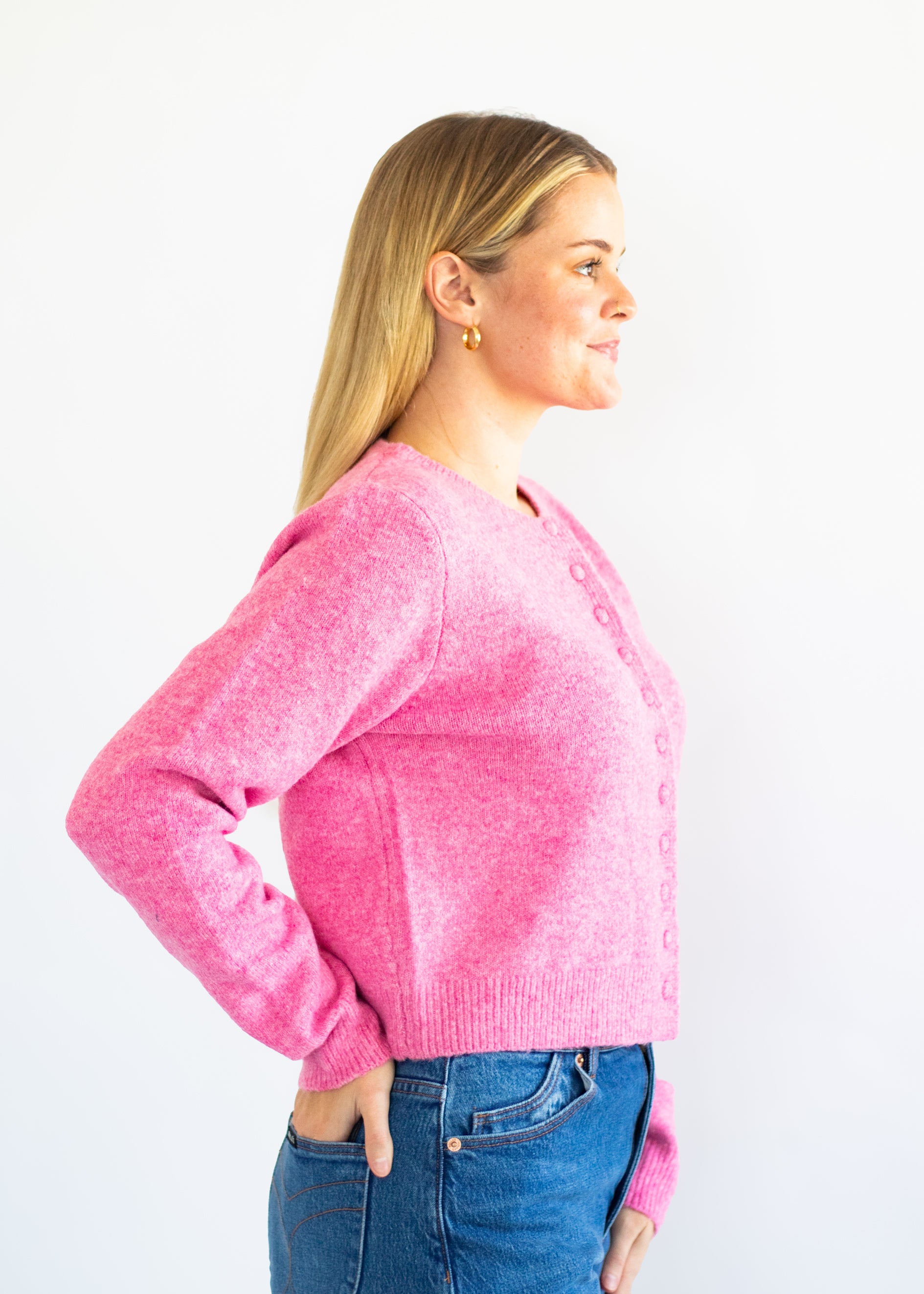 A woman in profile wearing a bright pink knit cardigan with buttons and blue jeans, standing against a plain white background.
