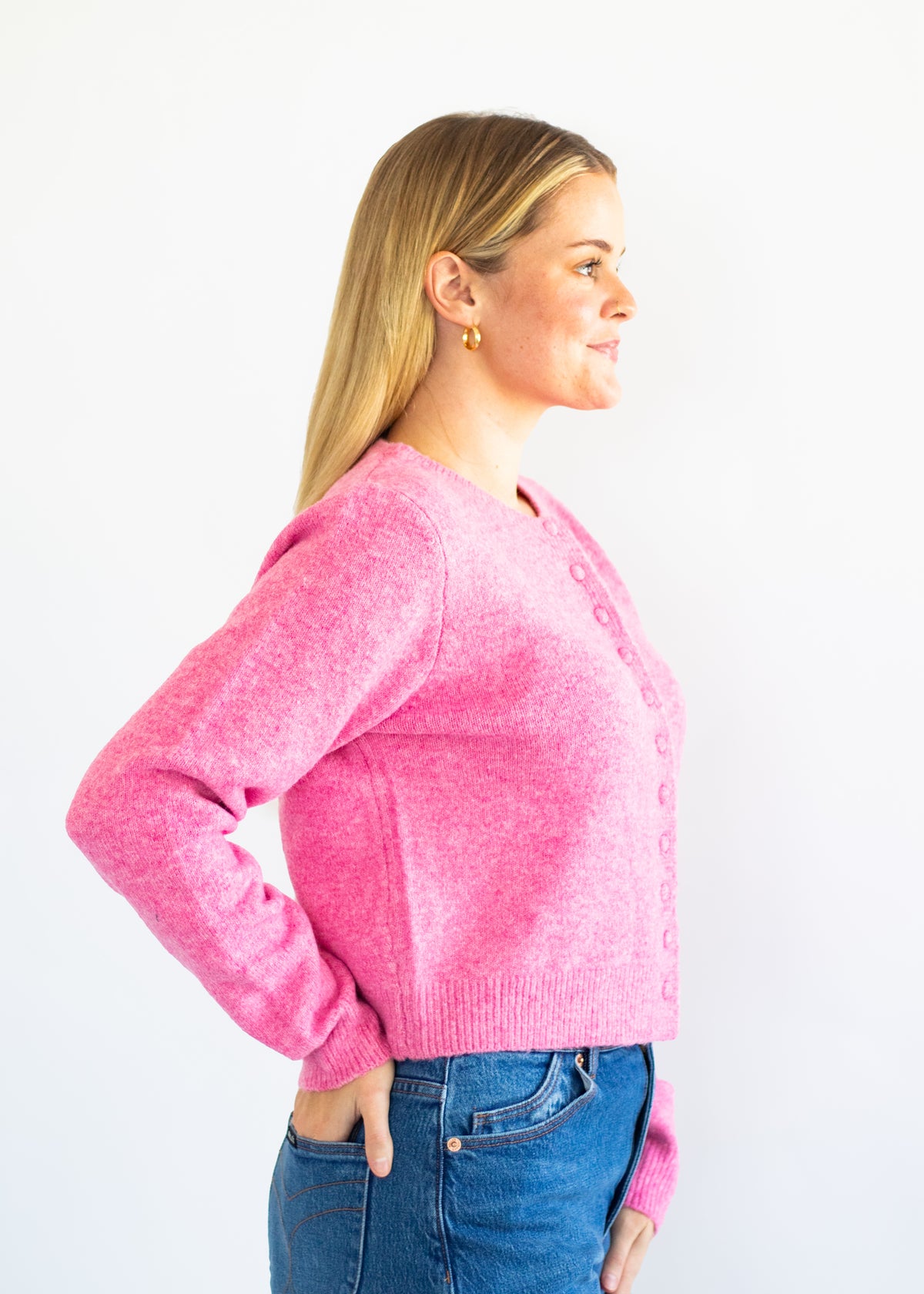 A woman in profile wearing a bright pink knit cardigan with buttons and blue jeans, standing against a plain white background.