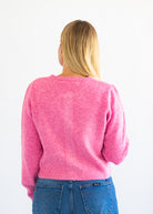 A woman viewed from behind wearing a bright pink knit sweater and blue jeans against a plain white background.