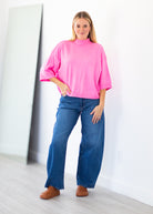 A woman modeling a bright pink mock-neck short-sleeve top with wide legs and blue jeans, standing in a minimalist room with a white wall and light wood floor.