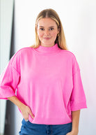 A woman wearing a bright pink mock-neck knit sweater with three-quarter sleeves and blue jeans, standing against a white wall.