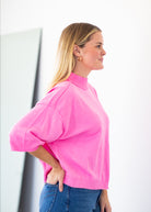 A woman in profile wearing a bright pink mock-neck knit sweater with voluminous elbow-length sleeves and blue jeans, standing against a clean white background.