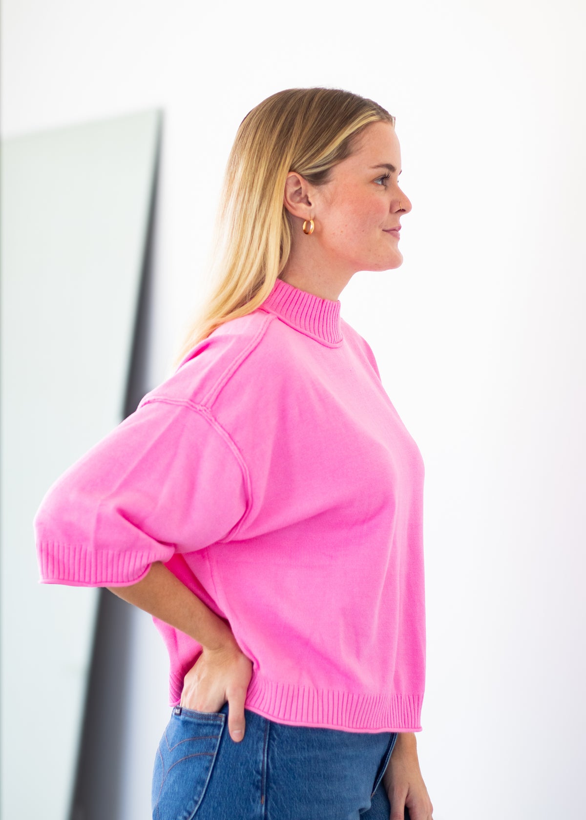 A woman in profile wearing a bright pink mock-neck knit sweater with voluminous elbow-length sleeves and blue jeans, standing against a clean white background.