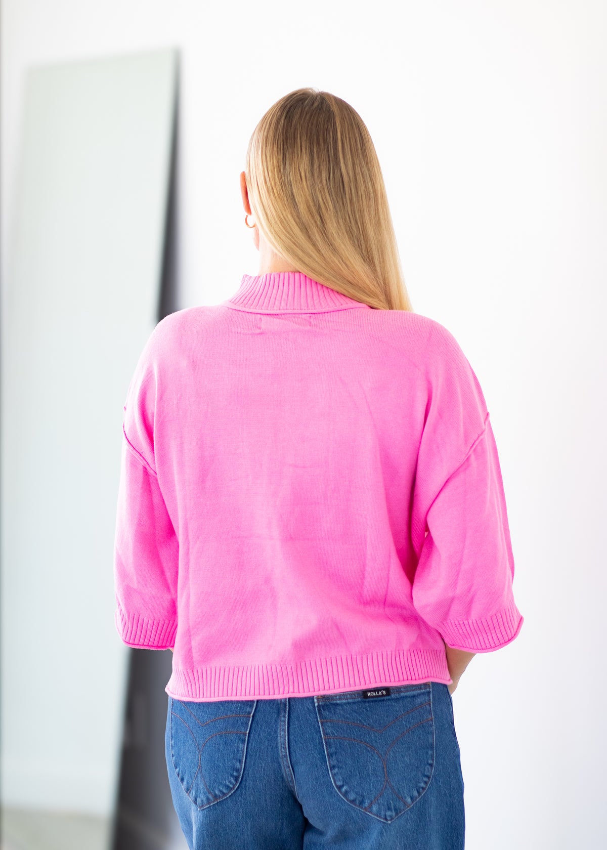 A woman viewed from behind wearing a bright pink knit sweater and blue jeans, standing in a minimalist indoor setting with a white wall and a tall mirror.