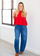 A woman modeling a bright red sleeveless peplum top with blue wide-leg jeans and brown shoes, standing in a bright room with a white wall and window.
