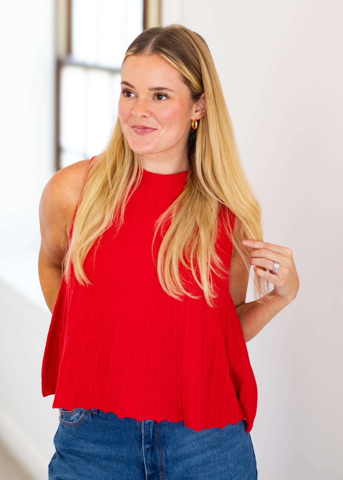 A blonde woman wearing a bright red sleeveless mock-neck top and blue jeans, standing in a brightly lit indoor setting.