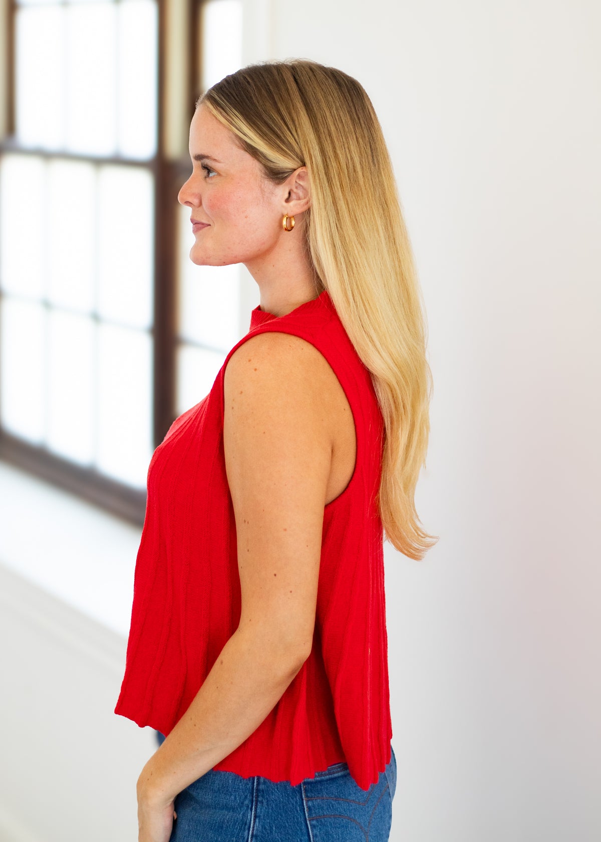 A woman with blonde hair is shown in profile wearing a bright red sleeveless top and blue jeans, standing indoors near a window.